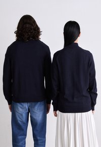 Due persone in piedi schiena contro schiena, indossano magliette a maniche lunghe nere; una con jeans blu e l'altra con una gonna plissettata bianca, su uno sfondo semplice.
