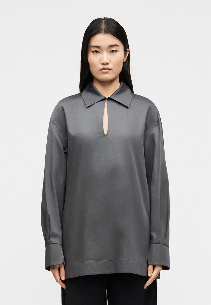 Γυναίκα με ίσια μαύρα μαλλιά φοράει υπερμεγέθη γκρι πουκάμισο με γιακά και μακριά μανίκια, με άνοιγμα κλειδιού στο μπροστινό μέρος, και μαύρο παντελόνι, σε λευκό φόντο.