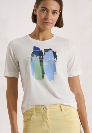 Femme aux cheveux courts et bruns portant un T-shirt blanc avec des coups de pinceau abstraits bleus et verts et un pantalon jaune.