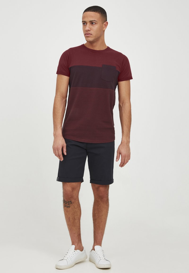 Maroon and dark blue striped t-shirt with a chest pocket, paired with navy shorts. Model wearing white sneakers, standing on a plain background.