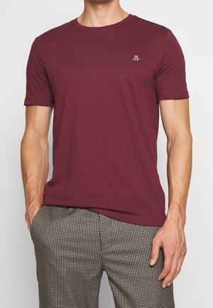 Mann trägt ein schlichtes burgunderfarbenes T-Shirt mit kurzen Ärmeln und kleinem weißen "M OP"-Logo auf der Brust und graue karierte Hose mit Händen in den Taschen.