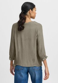Blusa leggera e texturizzata verde oliva con maniche 3/4 e polsini arricciati. Presenta bottoni decorativi sulla spalla. Indossata con jeans blu.