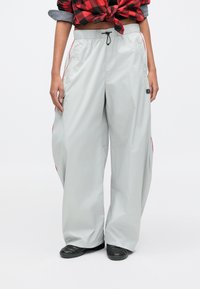 Jasnoszare spodnie o szerokich nogawkach z paskiem ściągającym w talii, z czerwonymi akcentami po bokach i naszywką z logo na lewej nogawce. Gładki, lekki materiał.
