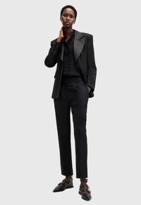 Svart skräddarsydd kostym med satinrevär, slim-fit byxor och en svart blus med spetsdetalj, i kombination med platta svarta skor.