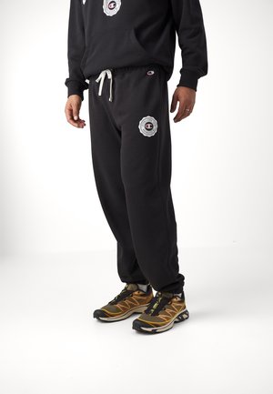 Person trägt schwarzen Champion-Kapuzenpullover und Jogginghose mit weißem Kordelzug und Logos, kombiniert mit braunen und schwarzen Sportschuhen.
