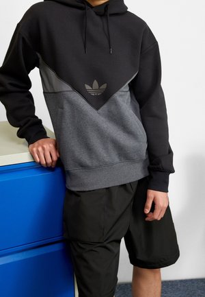 Persoon gekleed in een zwart en grijs Adidas hoodie met logo, zwarte shorts, leunend op een blauwe kast tegen een witte muur.