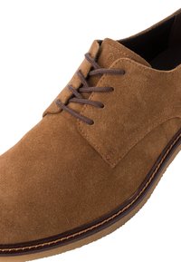Zapato de ante marrón con punta redonda, que presenta cordones marrones y un borde cosido sobre una suela de color beige. Textura suave y diseño minimalista.