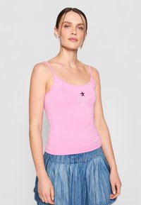 Rosa bomulls tanktop med tynne stropper, med en svart stjernegrafikk og "tommy girl" tekst, parret med et plissert denimskjørt.