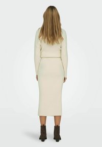 Vestido de punto acanalado en blanco roto, silueta ajustada, mangas largas, hasta la rodilla, combinado con botas de tobillo marrón oscuro.