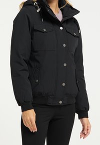 Giacca imbottita nera con cappuccio, caratterizzata da grandi tasche a patta con bottoni, polsini elasticizzati e coulisse al collo.