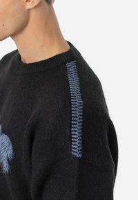 Suéter de lã preto com um decote redondo e um painel lateral com costura azul. A textura é suave e ligeiramente felpuda.