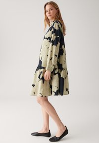 Frau, die vor einem einfarbigen Hintergrund in einem oversized blumenmuster Kleid in Schwarz und Beige mit langen Ärmeln und schwarzen flachen Schuhen steht.