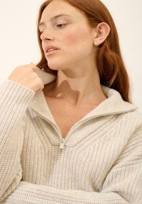 Maglione cremoso a trama con colletto alto e design a mezzo zip. Il materiale sembra morbido, con motivi a coste e a nido d'ape.