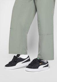 Czarne zamszowe sneakersy z białymi akcentami, okrągłym noskiem i grubą białą podeszwą; noszone z jasnoszarymi spodniami o szerokich nogawkach i białymi skarpetkami.