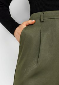 Olijfgroene broek met een gladde textuur, voorzien van een zijzak en riemlussen, gecombineerd met een aansluitende zwarte ribbed trui.