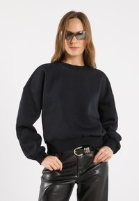 Felpa nera dal fit rilassato, con polsini a costine e scollatura ampia, abbinata a pantaloni in pelle nera a vita alta e occhiali da sole.