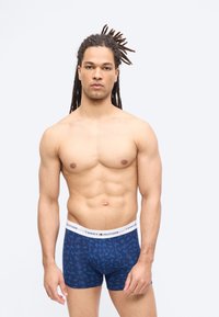 Άνδρας χωρίς πουκάμισο με μακριά dreadlocks, φορώντας μπλε μποξεράκια Tommy Hilfiger με σχέδια, στέκεται μπροστά σε απλό ανοιχτό φόντο.