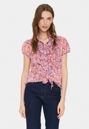 Floral print short-sleeve blouse in pink and blue with ruffled front detailing and a tied hem. Paired with dark blue jeans.
