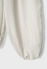Gros plan sur un pantalon en tissu beige clair avec un bas de jambe large et froncé, sur un fond uni.
