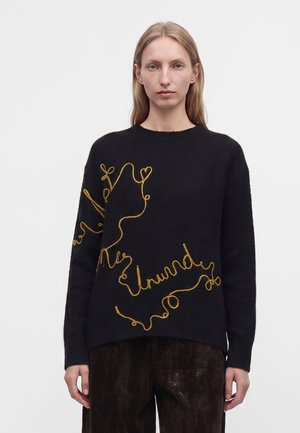 Kvinde med langt blond hår iført en sort sweater med gul kursivskrift og linjekunst, kombineret med mørkebrune bukser, på en hvid baggrund.