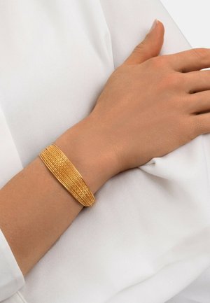 Bracelet en cuir texturé doré porté au poignet, présentant un design superposé avec une finition lisse et brillante sur un fond en tissu blanc.