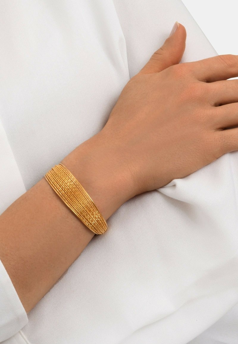 Bracelet en cuir texturé doré porté au poignet, présentant un design superposé avec une finition lisse et brillante sur un fond en tissu blanc.