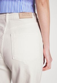 Jeans de denim de cor clara com um bolso traseiro, apresentando uma etiqueta de couro que diz "2NDDAY THINKTWICE" e um design de costura plana.