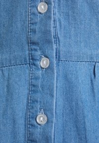 Blå denimtyg med en knäppdesign. Har ljusa sömmar och genomskinliga plastknappar. Slät textur med mjuk yta.