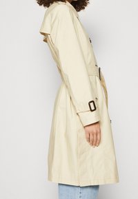 Beige trenssitakki, jossa on vyötärönauha, nappidekoraatio ja kevyt kiilto. Takissa on leveät kaulukset ja polvenmittainen siluetti.