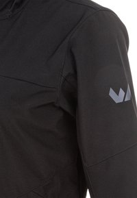 Manche de veste noire avec un petit logo géométrique réfléchissant sur le haut du bras et des coutures visibles.