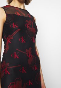 Black sleeveless dress featuring a sheer neckline and a red floral pattern with CK branding throughout. Smooth fabric texture.