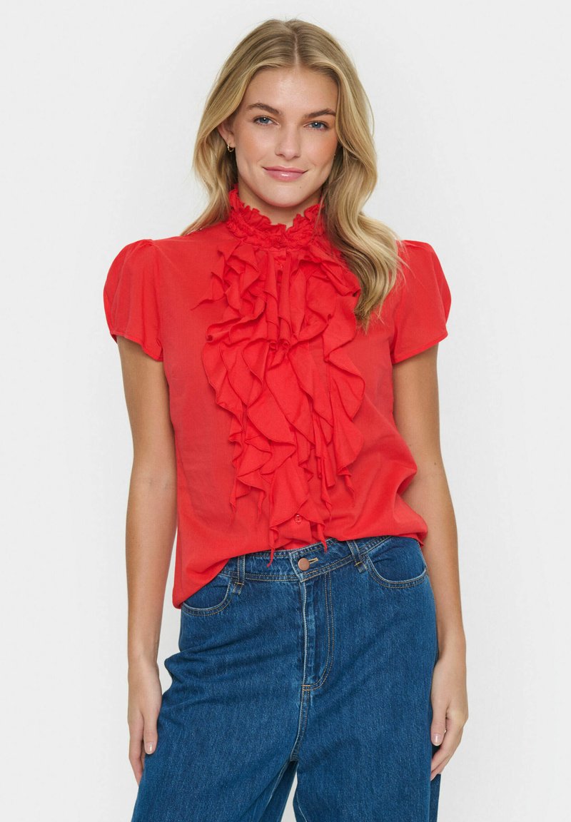 Blusa rossa con colletto arricciato e design frontale, maniche a sbuffo corte, realizzata in tessuto leggero, abbinata a jeans blu a vita alta.