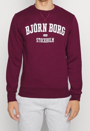 Bordowa bluza z białym napisem "BJÖRN BORG 1956 STOCKHOLM" noszona z jasnoszarymi spodniami dresowymi.