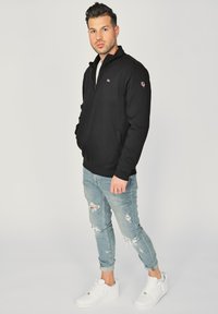 Czarna bluza z zamkiem błyskawicznym, z bocznymi kieszeniami, połączona z jasnoniebieskimi dżinsami z przetarciami i białymi trampkami. Gładka tekstura i luźny krój.