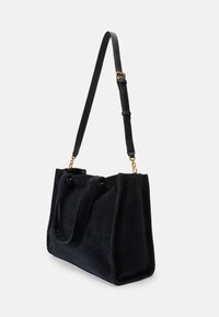 Saco tote de veludo cotelê preto com um formato plano, alça de ombro em couro, ferragens douradas e duas alças superiores. Tecido texturizado com riscas verticais.