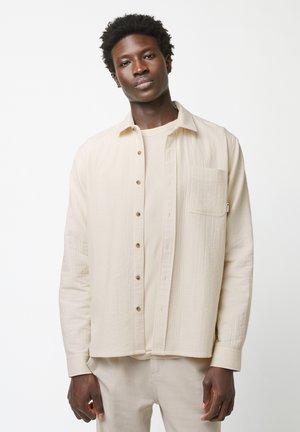 Beige textured shirt with buttons and a chest pocket, layered over a matching beige t-shirt. Model is wearing light beige pants.