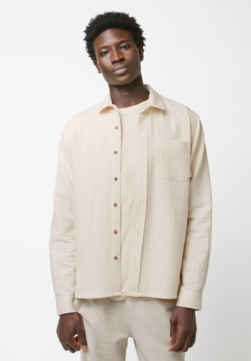 Beige textuur shirt met knopen en een borstzak, gelaagd over een bijpassend beige t-shirt. Model draagt lichtbeige broeken.