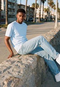 Ung mann i hvit t-skjorte og lyse jeans som sitter på en steinmur langs en solfylt bysti omgitt av palmer.