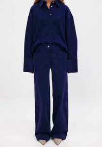 Tamsiai mėlynas kordūrinis marškiniai ir plačių kojų kelnės, su klasikiniu apykakle, mygtukų uždarymu ir tekstūriniu audiniu. Suderinta su smailiomis bežė batų kulnais.