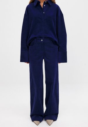 Ensemble en velours côtelé bleu marine comprenant une chemise à col classique et un pantalon à jambe large, avec une fermeture à boutons et un tissu texturé. Assorti à des escarpins beiges à bout pointu.
