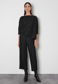 Schwarzes Strickoutfit mit einem locker sitzenden Oberteil und weit geschnittenen Hosen, die einen Schimmer und subtilen Glitzer haben. Knöchelhohe schwarze Stiefel runden den Look ab.