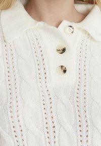 Weißer, gestrickter Pullover mit Zopfmuster, ausgestattet mit einem Kragen und drei goldenen Knöpfen entlang der Knopfleiste.