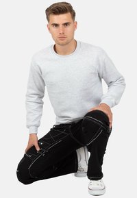 Felpa grigia con colletto rotondo, pantaloni cargo neri con cuciture bianche e sneakers bianche. Posa in ginocchio sollevando un ginocchio. Sfondo semplice.
