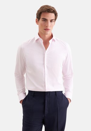 Jeune homme portant une chemise habillée rose clair à rayures rentrée dans un pantalon bleu marine foncé, mains dans les poches, sur un fond uni.