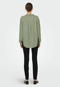 Lichtgroen shirt met lange mouwen en een afgeronde zoom, met ingebouwde knopen bij de manchetten en een detail op de rug, gecombineerd met zwarte slim-fit broek en platte schoenen.