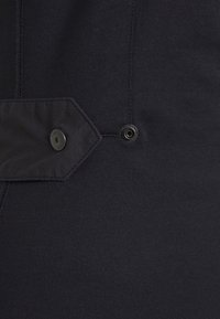 Tissu noir à la texture lisse, doté d'un rabat hexagonal avec fermeture par bouton. Un petit œillet est visible près du rabat.