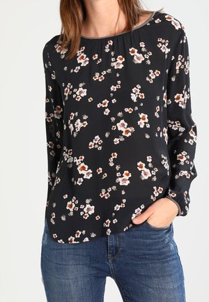 Blouse noire à manches longues avec un col arrondi, présentant un motif floral en blanc et rouille, fabriquée en tissu léger. Portée avec du denim.