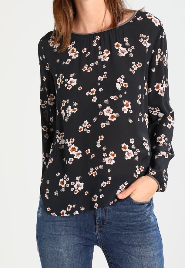 Blouse noire à manches longues avec un col arrondi, présentant un motif floral en blanc et rouille, fabriquée en tissu léger. Portée avec du denim.