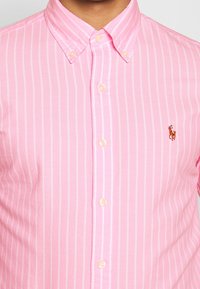 Ljusrosa skjorta med tunna vita vertikala ränder. Knapp-down krage, framplacket med ljusa knappar. Har en liten broderad logotyp.