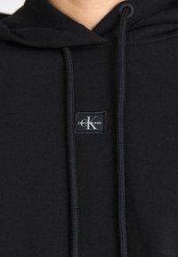 Juodas medvilnės džemperis su kišenėle priekyje, su raišteliais reguliuojama kapuce ir etiketė su "Calvin Klein Jeans" logotipu. Minkšta tekstūra ir vientisa spalva.
